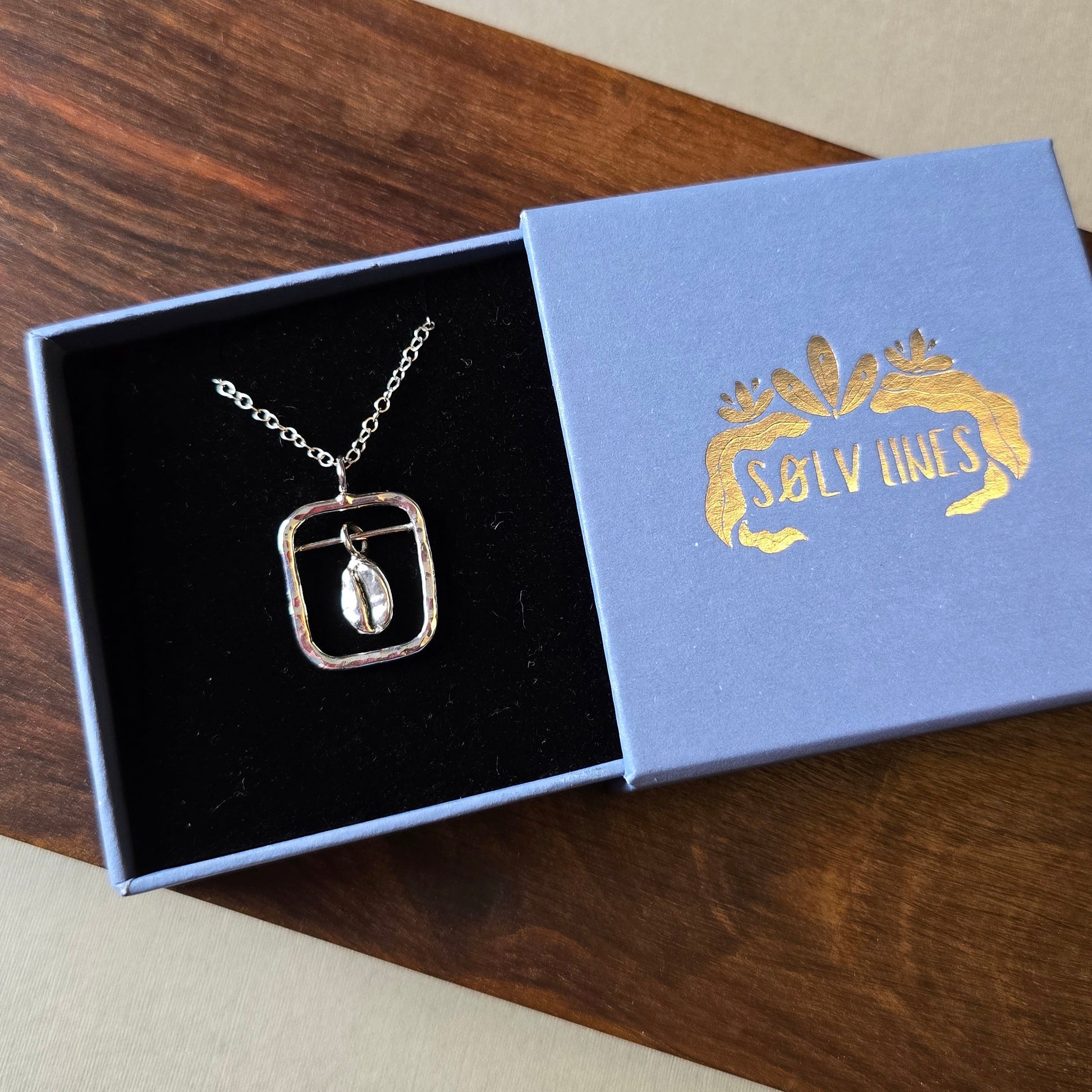 A sterling silver coffee themed necklace with a square silver frame around the bean in a jewellery box